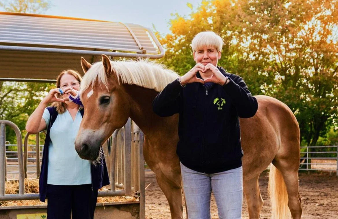 Zwei Mitarbeiterinnen formen mit den Händen ein Herz, dazwischen ein Haflinger auf der Koppel